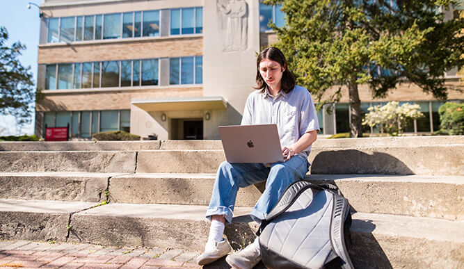 A focused college student sits on campus steps with a laptop and backpack, studying outside a modern academic building on a sunny day.