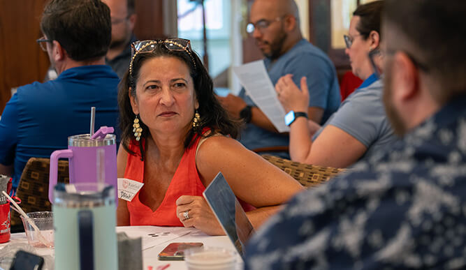 A female student in a red sleeveless top sits at a busy café table, listening thoughtfully amid people talking, reading papers, and sharing a lively conversation.
