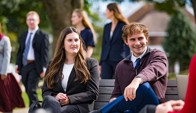 A smiling young man in a plum sweater and a woman in a black blazer sit on a bench outdoors, with students chatting in the background, creating a relaxed campus mood.