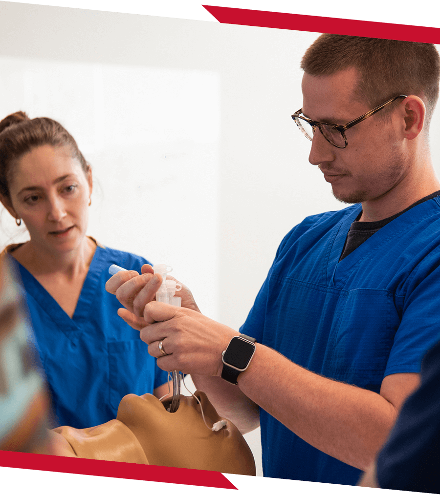 Students wearing blue scrubs practicing on a fake patient in a nursing class.