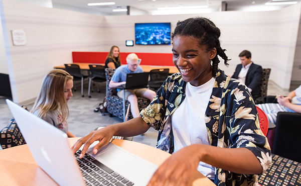 Female student at computer smiling with fellow students in the back. 