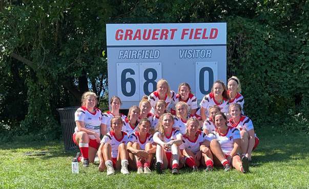 Cheerful members of the 2021-22 Women's Rugby Club sitting and posing in front of the Grauert Field scoreboard.