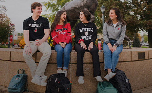 Four students in Fairfield attire, seated before the Stag statue on campus.