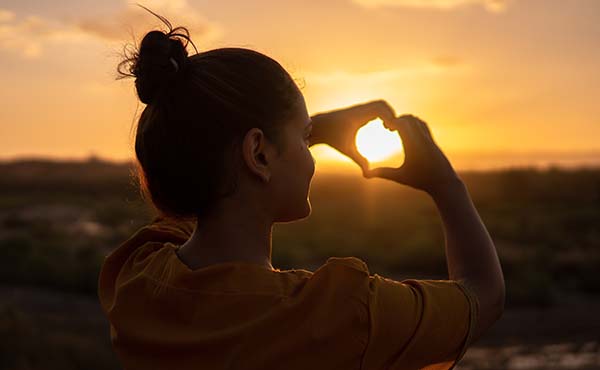 Image of a young woman holding her hands in the shape of a heart against the sunset.